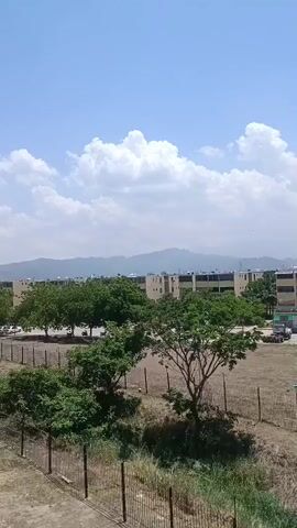 The video shows a daytime outdoor shot of a residential area under a partly cloudy sky. In the foreground, there's a metal fence, and beyond it, some vegetation. Further back, several trees with green foliage are visible, partially obscuring a row of multi-story apartment buildings. Mountains can be seen in the distant background, partially obscured by a haze. The buildings appear to be light-colored with some sections possibly painted green. Satellite dishes are visible on the roofs of some buildings.