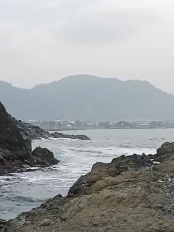 A video displays a coastal scene with rocky outcroppings in the foreground. The water is choppy with waves breaking against the rocks. In the background, a hazy mountainous landscape is visible, with a small town or settlement nestled along the coastline. The sky is overcast, creating a muted, diffused light. The overall impression is one of a rugged and somewhat subdued natural environment.