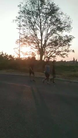The video shows a road with a large tree in the background. The sun is setting, casting a warm glow over the scene. Three people are running on the road. One person is wearing a dark shirt and shorts, another is wearing a blue tank top and shorts, and the third person is wearing dark clothing. They appear to be exercising or training. In the later part of the video, two people ride past on a motorcycle. The environment is rural, with vegetation and some structures visible in the distance.