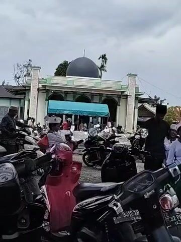 The video shows the exterior of a mosque. Many motorbikes are parked in front of the mosque. People, including some wearing white skullcaps, are moving around the motorbikes and towards the entrance of the mosque. The sky is overcast.