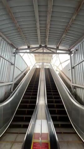 A low-angle shot shows two escalators moving upwards under a corrugated metal roof. The escalators have black steps with yellow indicator lines. The scene is illuminated by natural light coming from the end of the structure, creating a bright, washed-out effect. There are no people visible on the escalators. The environment appears to be a covered walkway or transit station.