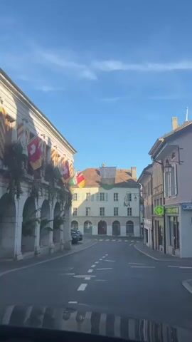 A wide shot captures a European town square under a clear blue sky. The perspective is from inside a vehicle, looking out through the windshield. On the left, a light-colored building with arched entrances and decorative flags displays a series of flags, including the Swiss flag. On the right, a street lined with buildings leads into the distance, with a green cross sign visible on one of the buildings, likely indicating a pharmacy. A banner with text hangs across the street between buildings in the center of the frame. The street is paved with asphalt and marked with white dashed lines. A few parked cars are visible in the square. The overall atmosphere is peaceful and quiet, possibly early morning or late afternoon, given the soft lighting.