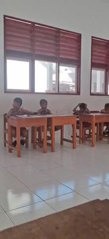 A classroom scene with several students sitting at desks. The room has white tiled floors and white walls. Windows with wooden blinds are visible on the left side. Several posters are hanging on the wall at the far end of the room. The students appear to be young and are dressed in school uniforms. Some are looking down at their desks, while others are looking around the room. The overall atmosphere is one of quiet concentration.