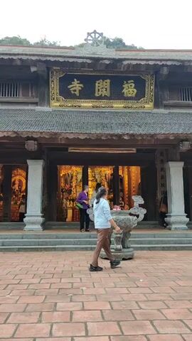 The video shows the front of a building with a large sign in Chinese characters. The building appears to be a temple or shrine with traditional East Asian architecture, including a tiered roof and stone columns. Several people can be seen entering or standing within the building. There is a stone incense burner in the foreground and a brick-paved courtyard. The background features a forested hill.