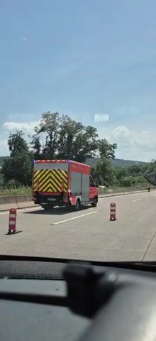 A red and yellow striped emergency vehicle, possibly a fire truck, is driving on a highway. The vehicle has the text "FEUERWEHR GERSHEIM" on its side. The scene is set on a sunny day with a clear blue sky and some scattered clouds. Green trees and vegetation line the side of the highway, and a bridge can be seen in the distance. There are traffic cones on the road, indicating some sort of closure or hazard. Another car, a dark hatchback with a license plate reading "37-XVS-5", is visible in the right lane. A person in an orange vest is standing on the side of the highway in the distance, possibly directing traffic or attending to an incident. The overall impression is one of an ongoing event on the highway.