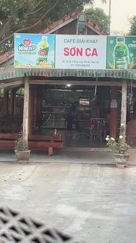 The video shows a roadside cafe called "Son Ca" in Vietnam. The cafe is a wooden structure with a green corrugated iron roof and a sign advertising a Manuka honey drink. A man is seen inside the cafe, possibly working. Outside, there are wooden benches and tables. A woman is sitting on one of the benches, using her phone. The area is paved with concrete, and there are trees and greenery around the cafe. In the distance, mountains can be seen. The overall impression is of a quiet, rural setting.