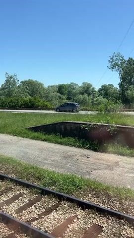 The scene presents a view from a moving train, with the focus on the railroad tracks and surrounding landscape. A concrete structure with overgrown grass borders the tracks. Adjacent to this is a road where several cars pass by. The background is filled with lush green trees under a clear, blue sky.