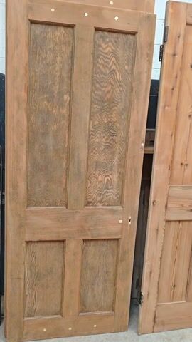The camera slowly pans right across a row of reclaimed wooden doors. The first door is a four-panel door with a unique grain pattern. The second door is a lighter wood with a two-panel design and a metal latch.  Several more doors are lined up behind this second door. The doors are leaning against a wall in a warehouse or workshop setting.  The lighting is bright.