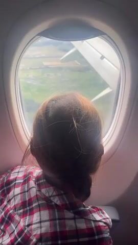 A young girl with braided auburn hair, wearing a red, white, and black plaid shirt, is seated next to an airplane window, looking out at the landscape below. The airplane's wing is visible outside the window, with the ground appearing green and somewhat rural. The girl is positioned so her back is to the camera.