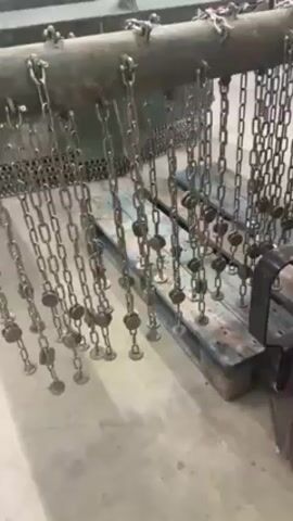 The video shows a piece of heavy machinery. It's painted in an olive-green color and appears to be some type of industrial equipment. Chains hang from a horizontal bar, each with a small, circular plate attached at the end. The chains are suspended over a wooden pallet. The background is an industrial-looking setting, with a concrete floor and various pieces of equipment visible. The camera moves around the machinery, showing different angles and details.