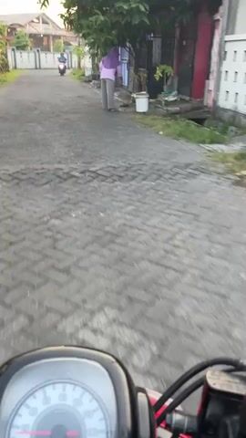 The video shows a POV shot from a motorcycle as it travels down a paved road. A person wearing a purple hijab and grey pants is walking away from the camera, towards a house on the right side of the road. A motorcycle is visible in the distance, approaching the camera. The environment appears to be a residential area with houses and vegetation visible. The time of day seems to be morning or evening, given the lighting conditions.