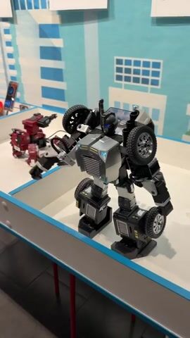 A static mid-shot captures a robot standing in a white tray with a blue border, situated on a red-legged table. The robot, primarily in shades of gray and black, boasts a distinct appearance with tires on its limbs and shoulders. Its chest is silver, while its head has a black visor-like structure. In the background, a blue wall features a white geometric pattern resembling buildings, hinting at a cityscape. To the left of the robot is a smaller, red robot with a gun-like appendage. A smartphone is visible in the far background, slightly to the left of the red robot. The background wall also displays written text, likely informational labels about the robots.
