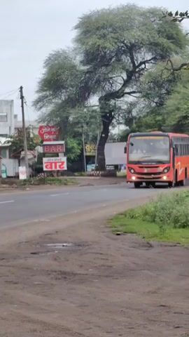 A red passenger bus travels on a paved road. The bus is marked with a white curved design and a logo on the side. A person is visible through one of the bus windows. The road has a dirt shoulder with grass and some puddles. In the background, there are trees, buildings, and advertising signs. The sky is overcast.