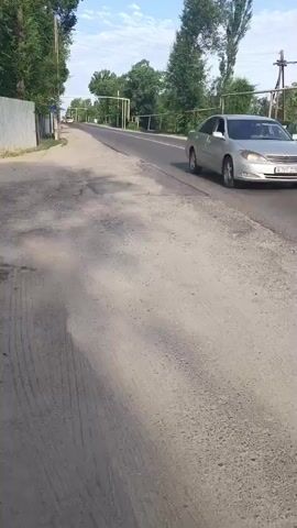 The video shows a paved road in a rural area. On the left side of the road is a dirt shoulder and a metal fence. On the right side of the road is a silver car driving away from the camera. The road is lined with trees and utility poles. The sky is blue with some clouds.