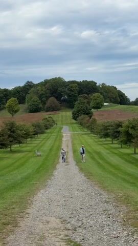 Two people walk along a gravel path in the middle of a wide, manicured lawn. The path stretches into the distance, flanked by rows of trees on either side. The lawn itself is neatly mowed in stripes. In the background, a wooded hill rises, with a building visible at its peak. The sky is cloudy.