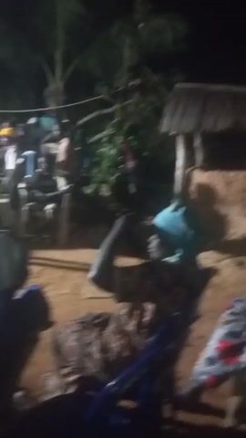 A grainy, dark, and shaky video shows a group of people gathered outdoors at night. They are sitting in chairs, and some are standing. The source of light is not apparent, but there is a bright light that creates long shadows. The ground appears to be dirt or sand. Some trees and a building can be seen in the background. Music is playing.
