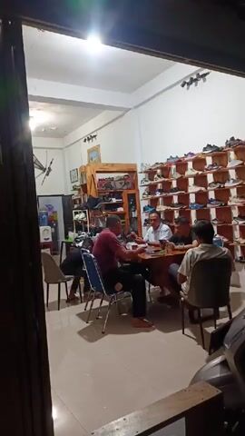 The video captures an indoor scene, likely a shoe store or a shop that sells footwear, in Bireuenmeunasahcapa, Indonesia, during the evening or night, indicated by the reliance on artificial lighting. The room features white walls, light-colored tiled flooring, and is illuminated by a bright overhead light.

Approximately five to six men are present. Four men are seated around a square, dark wooden table in the middle of the room, engaged in conversation. One man in a light blue collared shirt is actively speaking and gesturing towards the table. Another man, wearing a red shirt and dark trousers, sits facing him. Two other men are seated on the opposite side of the table. Further back in the room, another person is partially visible sitting at a smaller table or counter, and one more individual is partially obscured.

The right wall of the shop is lined with multi-tiered wooden shelves displaying numerous pairs of shoes, suggesting the primary business of the establishment. A larger wooden shelving unit is also visible in the background on the left side of the room, potentially holding more merchandise. Some bottles or containers are visible on a lower shelf in the background. The men appear relaxed, conversing in a casual setting. A motorcycle is partially visible outside the shop entrance on the right. No discernible text is visible within the frame.