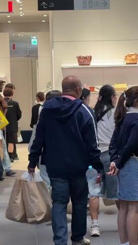 The video shows a group of people inside a store. In the foreground, a man wearing a dark blue hoodie and jeans is walking away from the camera, carrying two brown paper shopping bags in his left hand and a clear plastic bottle in his right. He is bald and has a pink collar visible on his shirt. To his right, two young women are standing and looking into a display window. The woman on the left is wearing a dark blue sweater and a denim skirt. The woman on the right has long dark hair and is wearing a light-colored jacket. Behind the bald man and slightly to the left, a man wearing a brown short-sleeved shirt and a baseball cap is walking away from the camera, carrying a brown paper shopping bag. In the background, other shoppers and displays of merchandise are visible. The store appears to be a boutique or department store, with light-colored walls and shelves displaying bags and other items. The lighting is bright and uniform. The overall scene suggests a commercial environment where people are shopping.