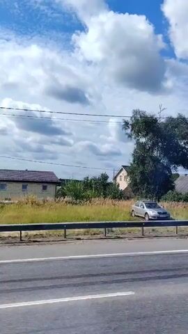 A white car is parked on the side of a road. Beyond the car, there is a tall tree with dense foliage, some houses with pitched roofs, and a dark fence. The sky is blue with scattered white clouds. A road with white lane markings is in the foreground. The video appears to be taken from a moving vehicle.