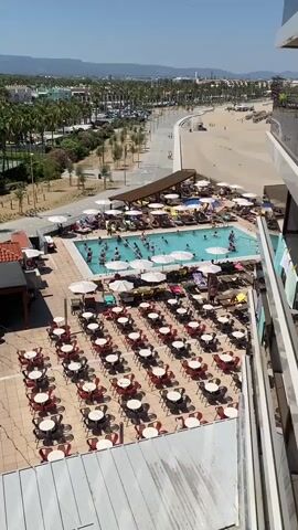 This video provides a high-angle, static view of an outdoor swimming pool area and a beach. The pool is filled with people swimming and sunbathing. Around the pool, there are numerous lounge chairs and umbrellas, with some people seated on the chairs. In the foreground, there is a patio with many tables and chairs, most of which are empty. Beyond the pool and patio is a sandy beach with people scattered on it, and further in the background, there are palm trees and buildings. The sky is clear and blue, suggesting a sunny day. The overall scene depicts a resort or hotel area with recreational facilities.