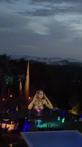 A nighttime aerial view shows a landscape with rolling hills and distant islands under a cloudy sky. In the foreground, a large, illuminated sculpture of a woman's bust is prominently displayed. She has light-colored skin, her hands are placed on her head, and she wears a patterned garment. To her left stands a tall, conical tower with lights. The ground below the sculpture is lit with various colored lights, including blue and green, and several small figures are visible in the distance, suggesting an event or gathering. A swimming pool is partially visible in the bottom right corner of the frame. The overall scene suggests an outdoor nighttime performance or art installation.
