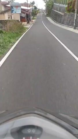 This first-person perspective video captures a motorbike ride along a paved road in Ketenger, Indonesia. The scene is set during the daytime, with overcast or cloudy weather, creating a diffused light. The road is asphalt with clear white lane markings, indicating it's a well-maintained thoroughfare.

On the left side of the road, there are several buildings. One building prominently displays a sign with text that appears to read "POJOKSARI" and "SIGN". Other buildings are visible further down the road, some with reddish roofs and what looks like an awning. There are also some plants and a few orange buckets placed along the sidewalk.

On the right side of the road, a stone wall borders a slightly elevated area, possibly a hillside or embankment. Vegetation, including trees and bushes, can be seen growing along this embankment.

Several people are visible riding motorbikes on the road ahead, suggesting it's a common mode of transportation in the area. The rider's motorbike dashboard is partially visible at the bottom of the frame, showing some indicators and a dial that might represent speed.

The overall atmosphere appears calm and ordinary, depicting a typical journey on a rural or semi-urban road in Indonesia. There are no strong emotional cues, but the video conveys a sense of movement and travel through the landscape.