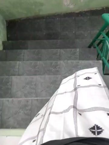 A first-person perspective video shows someone walking down a staircase. The person is wearing a white sarong with a black checkered pattern. The staircase has dark gray tiled steps and a green metal railing. The walls are painted a pale green and show signs of wear and tear. The lighting appears to be natural, possibly from an overhead source. The video is shaky and appears to be recorded on a mobile device.