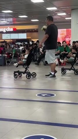 This media file shows a man in a black t-shirt and light grey shorts pushing two children in separate strollers through an airport. The man is walking towards the right of the frame. He is carrying a bag of items in his left hand. The children appear to be young, and they are both sitting in lightweight, foldable strollers. One stroller is slightly in front of the other.

In the background, there are other people seated and standing. To the left, there is a sign for "Gong Cha," a beverage chain. The setting is a well-lit indoor area, likely an airport terminal, with a tiled floor and overhead fluorescent lighting. The floor has blue painted lines and circular directional arrows. A woman with a light blue shirt and black pants, carrying a light green tote bag, walks past the man and strollers from the right side of the frame towards the left. The overall scene suggests a travel day, possibly at an airport in Busan, South Korea, given the location context. The people in the background appear relaxed, and the man in the foreground seems focused on maneuvering the strollers. The time of day is not explicitly indicated, but the indoor lighting suggests it could be any time.