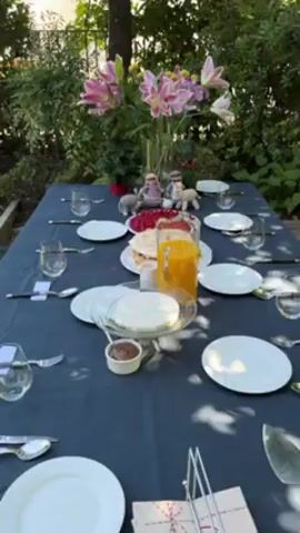 The video shows a neatly set table in a garden. The table is covered with a blue cloth and set for eight people, with white plates, cutlery, and glasses at each place setting. In the center of the table, there is a large vase filled with pink lilies. Several food items are arranged around the vase, including a pitcher of orange juice, a plate of raspberries, and a meringue dessert. There is also a small bowl of what appears to be chocolate spread and a dish of white shredded coconut. Between the lilies and food items, there are four small knitted figures, including two figures resembling people and two figures resembling animals. The garden is lush and green, with trees and bushes visible in the background.
