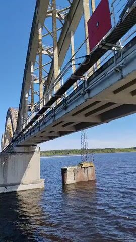 The camera moves beneath a long bridge over a body of water. The bridge is constructed of concrete and steel. The water is dark blue with ripples on the surface. In the background, a thin line of trees can be seen on the opposite shore. A number '5' is painted on one of the concrete bridge supports. To the right, an electrical tower stands on a platform in the water. The sky is a clear, bright blue. The wind is audible in the recording.