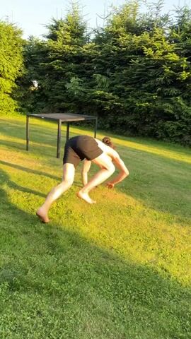 A person is on a grassy lawn in front of a dense green hedge. A dark rectangular table is to the left of the person. The person, wearing black shorts, is on all fours and appears to be crawling or moving their body in a way that involves their torso being very low to the ground. Their head is down and their legs are extended behind them. The person then moves their body in an arc, and appears to be going under the table. The person is barefoot.