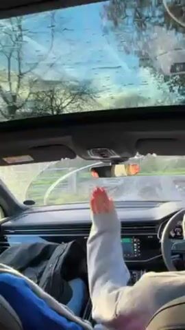The video shows the interior of a car with a panoramic sunroof. It’s raining, and the windshield wipers are in motion. A person in a white hoodie is driving, their right hand visible on the steering wheel. A jacket is on the passenger seat. The car is driving down a winding road lined with trees.
