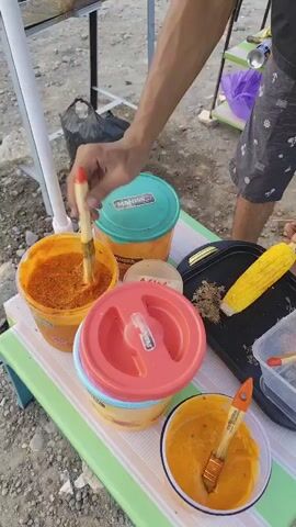 A person is brushing a piece of corn with a yellow sauce. There are three containers with lids, one green, one orange and one red, on a table covered with a striped cloth. Next to the corn is a grill pan and a small plastic container with brushes inside. In the background, a black trash bag is visible. The ground appears to be sandy or gravelly.