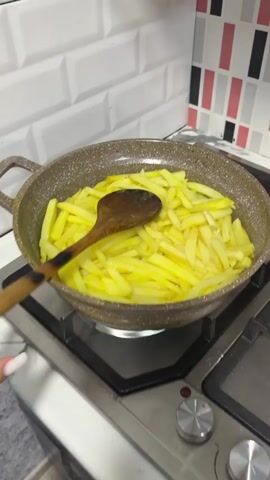 A pair of hands is stirring sliced potatoes in a pan with a wooden spoon. The pan is on a gas stove. The potatoes are yellow and appear to be frying in oil. The background is a white tiled wall with a mosaic backsplash.