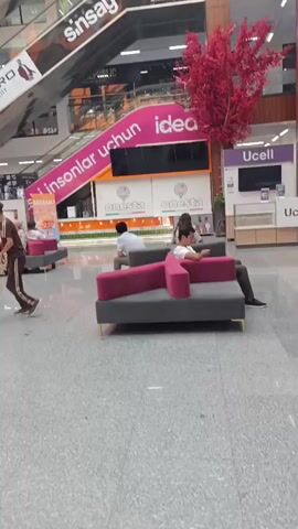 The video pans across the interior of a multi-story shopping mall. A bright, open atrium features escalators, shops with signage such as "sinsay" and "idea," and seating areas with magenta couches. Green and yellow vertical decorations hang from the ceiling, and a decorative red tree is visible. Several people are present, some walking and others seated, one person is taking a photo with their phone. The mall appears clean and well-lit, with a modern architectural design.
