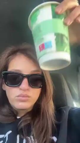 A woman wearing sunglasses is sitting in a car and shaking a cup over her head. The cup is white with a green design. The woman has brown hair. She is wearing a black sweatshirt with white writing on it. The car seat is dark. The inside of the car is visible in the background.