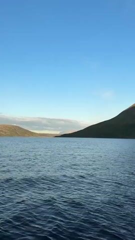 The video shows a scenic view of a body of water with mountains in the background. The water is calm with gentle ripples. The mountains are covered in green vegetation at the base and rocky terrain further up. Some parts of the mountain peaks are lit by sunlight, while others are in shadow, indicating the time of day might be late afternoon or early morning. The sky is clear and blue with a few wispy clouds near the horizon. The perspective suggests the footage is being filmed from a boat or a similar vantage point on the water, moving slowly or stationary. There are no people or discernible objects in the water or on the shore. The overall impression is one of natural beauty and tranquility.