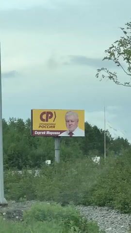A video captured from a vehicle shows a billboard featuring a man with white hair wearing a white shirt and red tie. The billboard displays text in Russian, including the words "СПРАВЕДЛИВАЯ РОССИЯ" which translates to "A Just Russia." Road signs are also visible: a yield sign (a yellow diamond with a white border) and a warning sign (a white triangle with a red border and a black symbol depicting a T-junction). Further down the road, a line of streetlights accompanies a lush green landscape. In the background, snow-capped mountains emerge from behind a dense forest, suggesting a mountainous and possibly cold climate. The sky is overcast with scattered clouds. The video appears to be filmed while the vehicle is in motion.