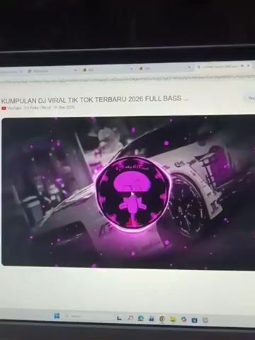 A video is playing on a laptop screen. The video is a YouTube video titled "KUMPULAN DJ VIRAL TIK TOK TERBARU 2026 FULL BASS". The video features a visualizer with a cartoon character in the center, which changes color from purple to blue to green. The background of the video is a graphic of a white sports car with purple accents. The YouTube channel name is "DJ Pinky Official". The video was uploaded on September 11, 2020. The laptop is open, and the screen is lit up. The surrounding environment is dark.