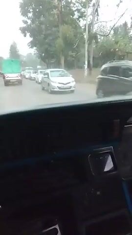 The video shows a view from inside a car, looking out onto a road. There is a line of cars and a truck ahead, all traveling in the same direction. The road is paved and flanked by trees and utility poles. The weather appears overcast. The camera remains relatively still inside the car, capturing the movement of the vehicles on the road ahead.