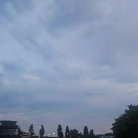 The video shows a time-lapse of a cloudy sky with a tree in the foreground in the first frame. The sky gradually changes, revealing hints of pink and orange as the sun sets in the second frame. The third and fourth frames show power lines and towers against the cloudy sky, with some buildings and a street visible in the distance. The final frame depicts a road with streetlights and power lines, as well as a parked truck, indicating an urban or industrial environment. The overall atmosphere is calm and transitions from daytime to dusk.