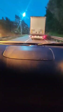 The video is filmed from the dashboard of a vehicle driving on a road at dusk or night. Ahead of the vehicle, a large white truck with a tan trailer is visible. The truck's brake lights are illuminated, indicating it is slowing down or stopped. The road appears to be a two-lane highway with trees lining the right side. A streetlight is visible in the distance on the left side of the road. The interior of the vehicle is dark, with the dashboard illuminated by the faint light from the exterior and the truck's lights. There are no people visible within the frame of the video.