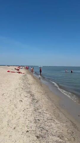 This video shows a serene beach scene in Rewa, Poland. The bright, clear blue sky suggests a sunny day, likely midday given the strong sunlight casting shadows. The water is a calm, clear turquoise, with small waves gently lapping at the sandy shore.

Several people are visible enjoying the beach and the water. On the left side of the frame, a few individuals are walking along the waterline, dressed in swimwear. Further out in the sea, a small group of people are wading or swimming, their bodies partially submerged in the water. One person appears to be floating or swimming more actively in the middle distance. In the far distance, a few sailboats are scattered across the horizon, adding to the tranquil maritime atmosphere.

The beach itself is composed of light-colored sand, with some scattered pebbles and seaweed along the tide line. The overall impression is one of a peaceful and relaxing summer day at the seaside. There is no visible text in the video.