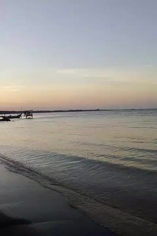 A vertical video shows a beach with dark sand and the ocean. The sky is a pale gradient of blue and pink, indicating either dawn or dusk. In the distance, there is a faint horizon line with structures that appear to be piers or small buildings. Two small boats are visible near the shore on the left side of the frame, and a small structure that looks like a raised platform or shack is in the water near the boats. Gentle waves are lapping at the shore. The overall mood is serene and peaceful.