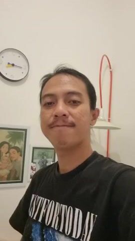 A man with dark hair and a mustache is looking at the camera. He is wearing a black t-shirt with white text that reads "UST FOR TODAY." Behind him, on the wall, is a framed picture of three people, and a round clock. To the right of the man is a white lampshade with a red cord. The man blinks and then lowers his head.