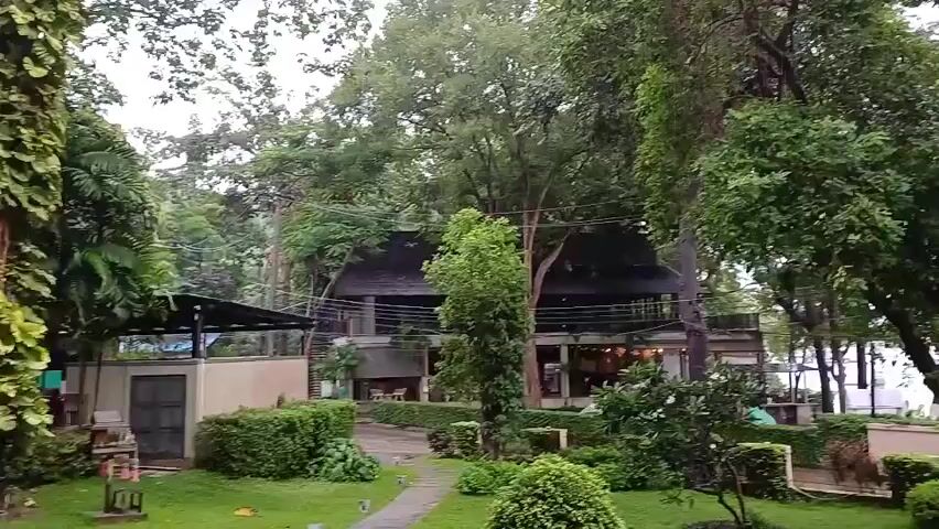The video pans across a lush green garden area with manicured bushes and trees. In the background, a multi-story building with a dark roof and balconies is visible, surrounded by more trees. Beyond the building and trees, a body of water, likely the ocean, can be seen with some structures, possibly boats or piers, and a playground set on the shore. The sky is overcast.