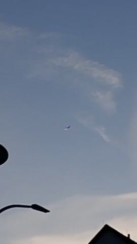 The video shows a clear blue sky with some wispy clouds. In the distance, a small object appears to be in flight, possibly an airplane. In the foreground, the silhouette of a street lamp and the roof of a building are visible against the sky. The lighting suggests it is either early morning or late evening. The object in the sky appears to be moving across the frame.