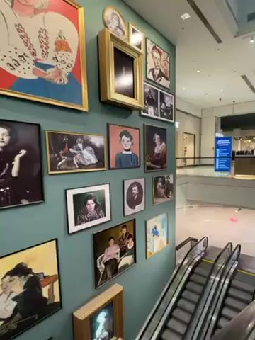 The camera pans up a wall covered in framed paintings and photographs of people, displayed above a set of escalators in what appears to be a commercial building or mall. The artwork varies in style and framing, creating a gallery-like effect. The wall is painted a light teal color. The escalators are silver and black, leading to a higher level visible in the background. A blue sign is also visible in the distance.