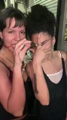 Two young women stand close together outdoors at night, possibly on a city street. The woman on the left has dark hair with bangs and is wearing a red and black top and a silver necklace. She holds a red beaded rosary in front of her face, biting the beads. The woman on the right has curly hair pulled up and is wearing a black tank top. She covers her face with one hand. She has a tattoo on her arm and a ring on her finger. A metal security gate and a light fixture are visible in the background.