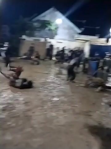 At night, an outdoor gathering takes place in a muddy, unpaved area in Jenggrik, Indonesia. Numerous people, predominantly adults dressed in dark, possibly black, attire, are present. In the foreground, one individual falls to the ground and quickly recovers, moving in a seemingly performative or ritualistic manner. Other participants are scattered around, some standing, some sitting, observing the activity.

The ground is saturated with mud and water, suggesting either recent rainfall or an intentional part of the event's setting. In the background, illuminated by artificial lights, are several simple houses or buildings. A wooden fence or barrier is visible in the mid-ground, and a blue barrel sits on the right side of the frame. The overall atmosphere appears engaged, possibly part of a cultural performance or communal activity. No discernible text is visible in the media.